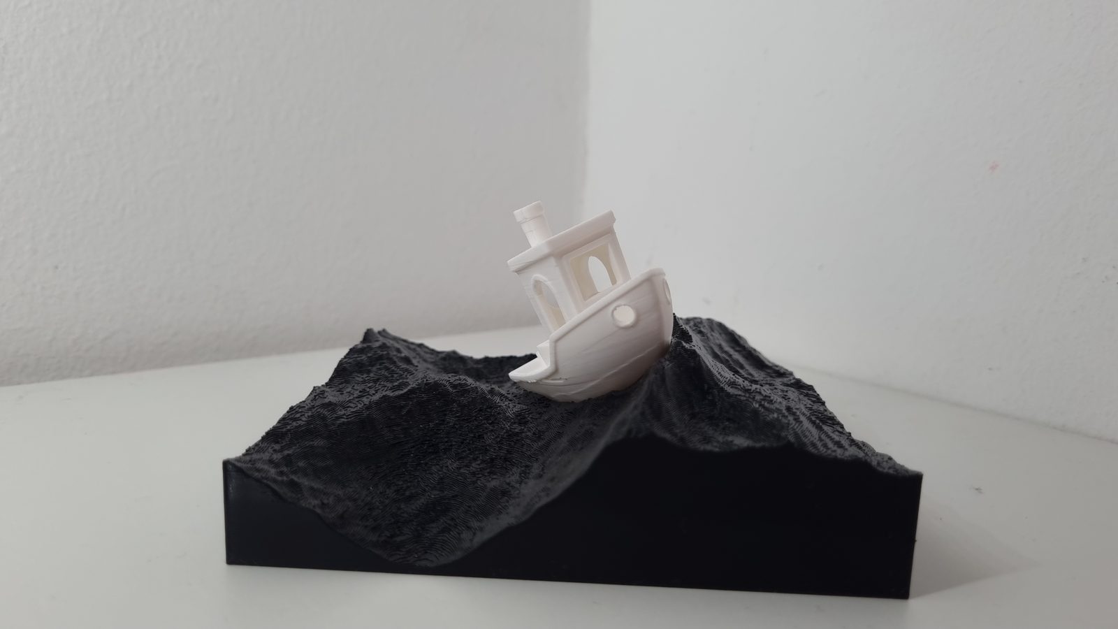 Weißes Schiffsmodell schräg im Wellental eines gedruckten schwarzen Reliefsockels, wirkt wie in bewegter See