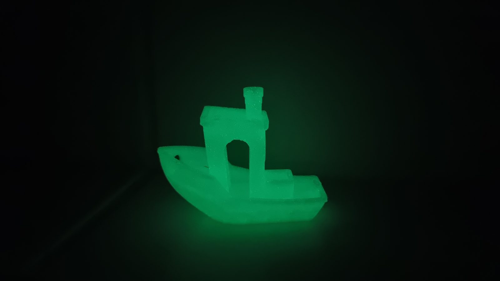 Ein Schiffsmodell aus nachleuchtendem Filament leuchtet im abgedunkelten Raum grün und wirft einen grünen Lichtschein auf die Unterlage