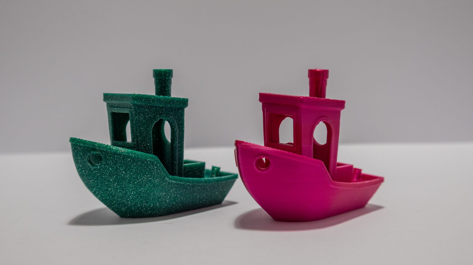 Zwei baugleiche Miniatur-Schiffsmodelle nebeneinander, links in glitzerndem Petrolgrün, rechts in kräftigem Magenta, vor neutral grauem Hintergrund