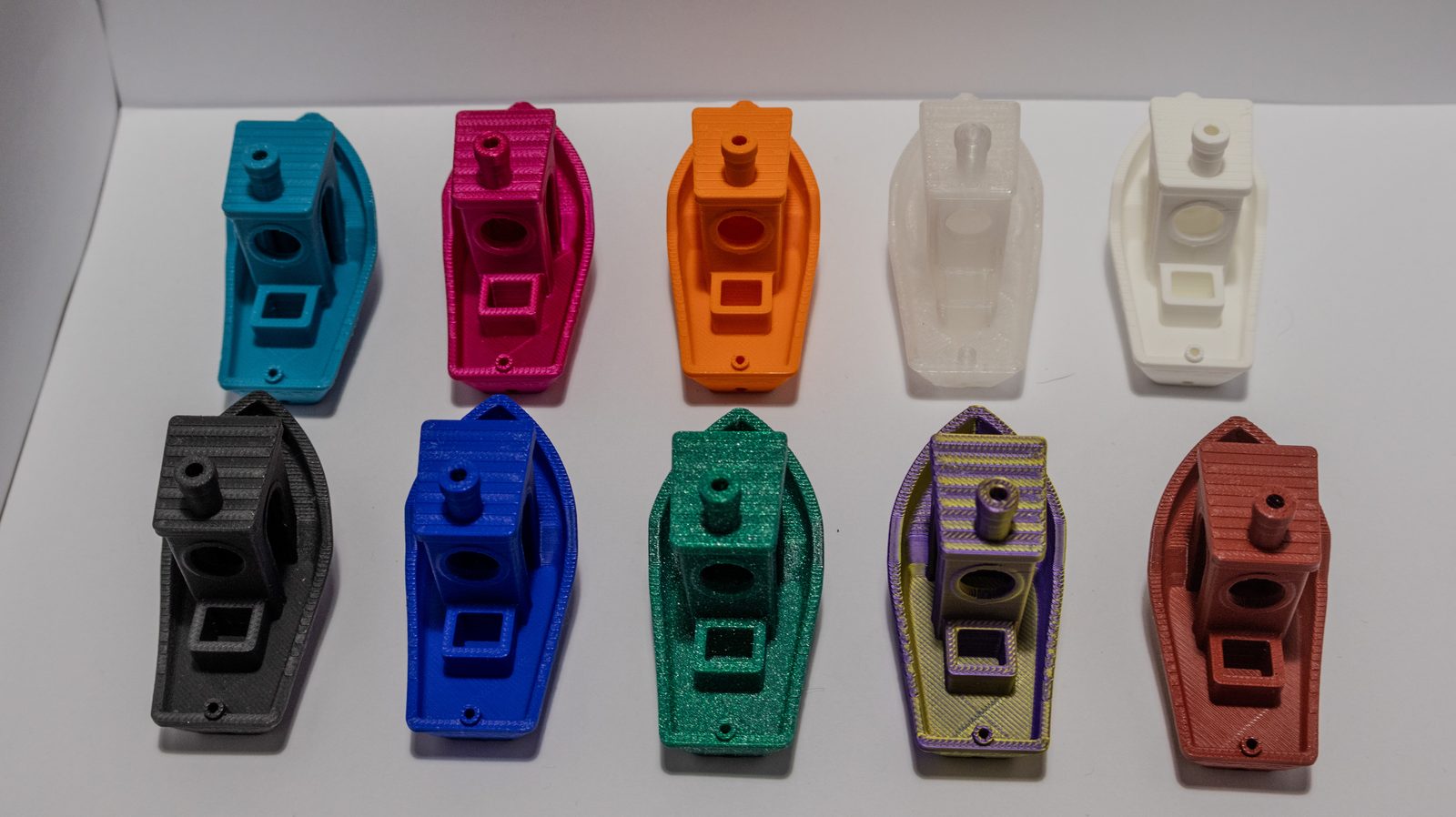 Zehn baugleiche Schiffsrumpf-Bauteile in zwei Reihen, jedes in einem anderen Filament — von Türkis über Transparent und Anthrazit bis zu Glitzergrün und Kupferrosa